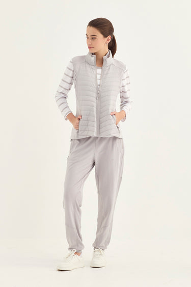 A woman modeling a light gray puffer vest over a white striped long-sleeve top, paired with matching light gray jogger pants by Verge, and white sneakers, against a plain white studio background.