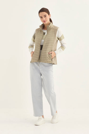 A woman modeling a sleeveless quilted puffer vest in a light beige/khaki color, styled over a white long-sleeve top and light gray wide-leg jeans by Verge, against a plain white studio background.