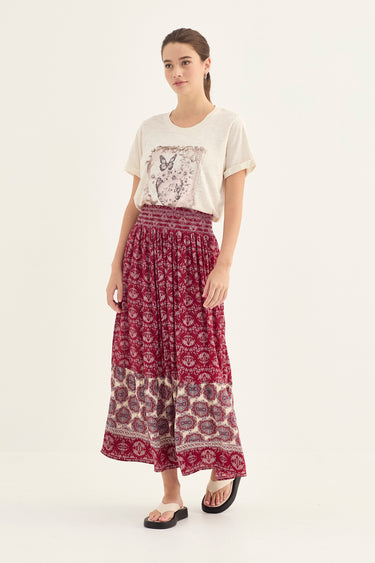 A woman modeling a bohemian-style outfit featuring a cream graphic t-shirt and a long, pleated maxi skirt by Verge with a deep magenta and beige paisley print.