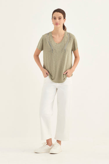 A woman modeling a short-sleeve V-neck top in a light sage green color with silver-gray embroidered detailing around the neckline, paired with white cropped pants by Verge and white sneakers, against a plain white studio background.