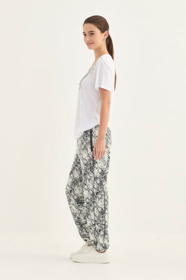 A woman modeling loose, wide-leg pants with a black and white abstract floral pattern by Verge, paired with a white t-shirt and white sneakers, standing against a plain white studio background.