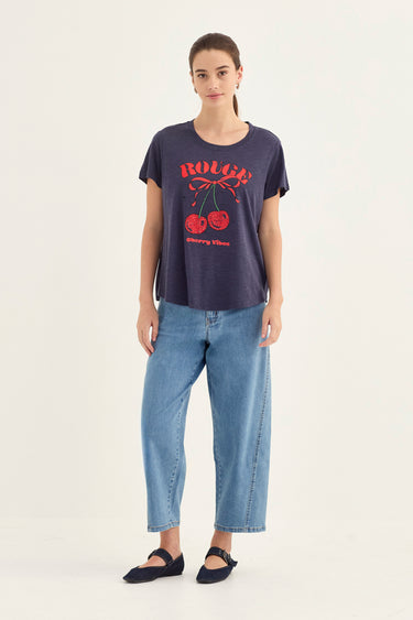 A woman modeling a dark navy blue graphic t-shirt with red cherry and text print, paired with light blue jeans by Verge and black flats, against a plain off-white studio background.