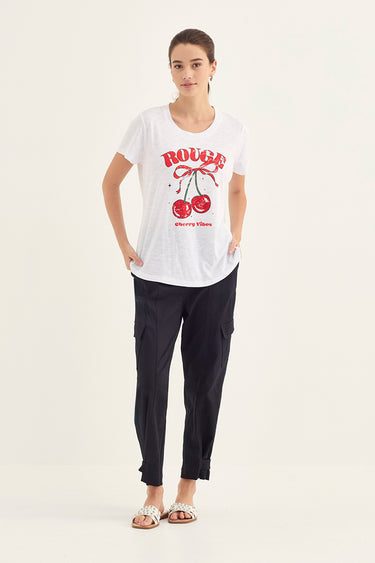 A woman modeling a white short-sleeve graphic T-shirt with a red 'ROUGH' text and cherry print, paired with dark cargo pants by Verge and metallic sandals, against a plain off-white studio background.