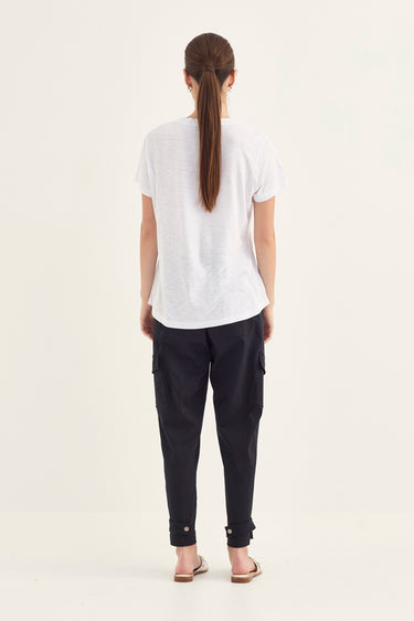A woman viewed from behind wearing a white short-sleeve t-shirt, dark navy cargo pants by Verge, and flat sandals, standing against a plain off-white studio background.