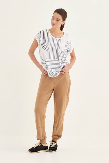 A woman modeling a white short-sleeve top with black geometric stripes and a gathered waist, paired with pants by Verge and black sneakers, against a plain light background.