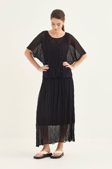A woman modeling a black pleated maxi top and skirt with sheer short sleeves and a layered, flowing silhouette by Verge, paired with black sandals, against a plain off-white studio background.