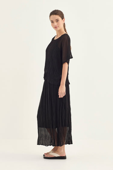 A woman modeling a black two-piece outfit consisting of a short-sleeve top and a pleated maxi skirt by Verge, photographed against a plain light background.