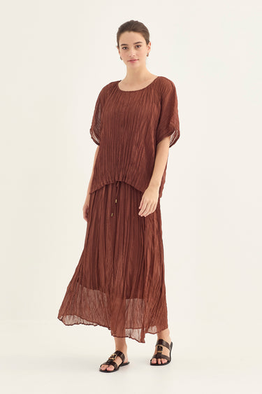 A woman modeling a brown pleated maxi top with flutter sleeves and a skirt with a drawstring waist by Verge, paired with black sandals, against a plain light background.