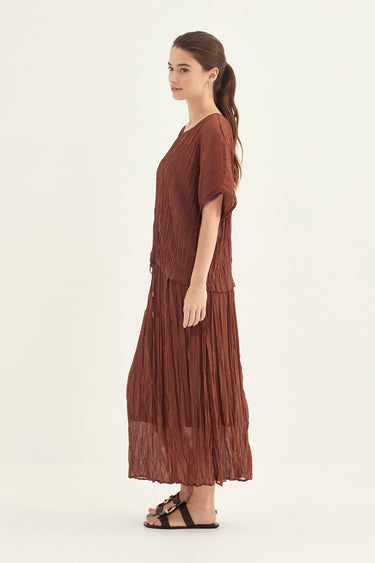 A woman modeling a brown pleated midi dress with short sleeves and a V-neckline by Verge, shown in profile against a plain light background.