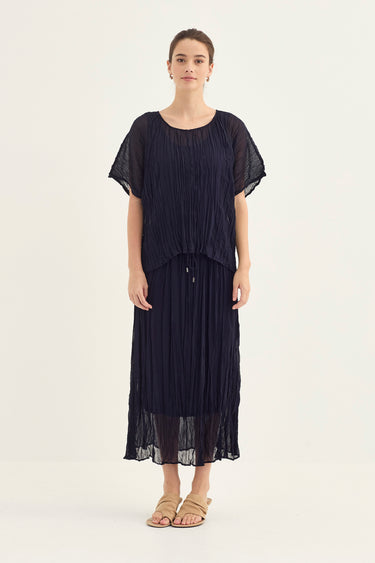 A woman modeling a dark navy blue pleated top and skirt with short flutter sleeves by Verge and a buttoned waist, paired with beige flat sandals, against a plain off-white studio background.