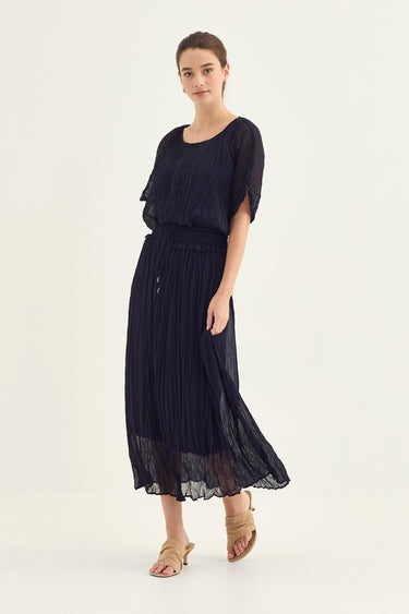 A woman modeling a dark navy blue midi dress with a pleated skirt and short flutter sleeves by Verge, paired with beige strappy sandals, against a plain light background.