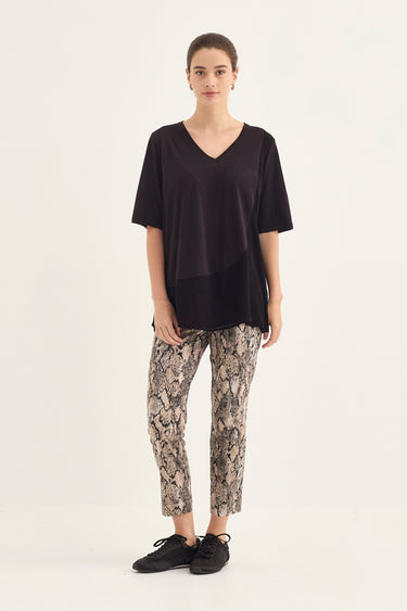 A woman modeling a black short-sleeve V-neck top and beige snakeskin print cropped pants by Verge with black sneakers, standing against a plain off-white studio background.