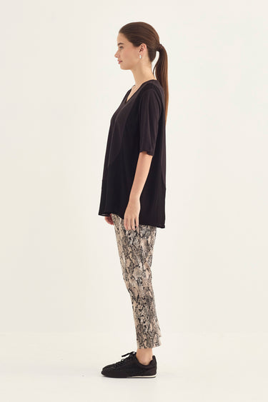 A woman shown in profile wearing a loose black short-sleeve top, beige and gray snakeskin print pants by Verge, and black slip-on shoes against a plain off-white studio background.