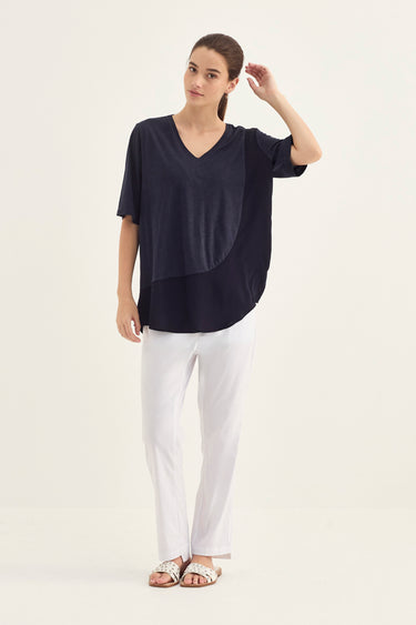 A woman modeling a short-sleeve V-neck top in dark navy with an asymmetrical draped panel detail, paired with white pants by Verge and metallic sandals, against a plain off-white studio background.