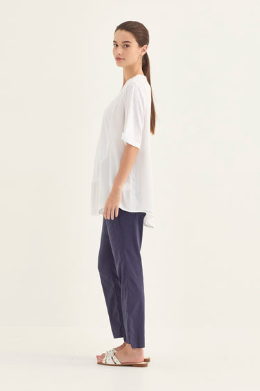A woman stands in profile wearing a loose white short-sleeve top, dark navy pants by Verge, and white slide sandals against a plain off-white studio background.