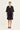 A woman stands against a plain light background wearing a black short-sleeve blazer over a black button-front top and black shorts by Verge, with black strappy flat sandals.