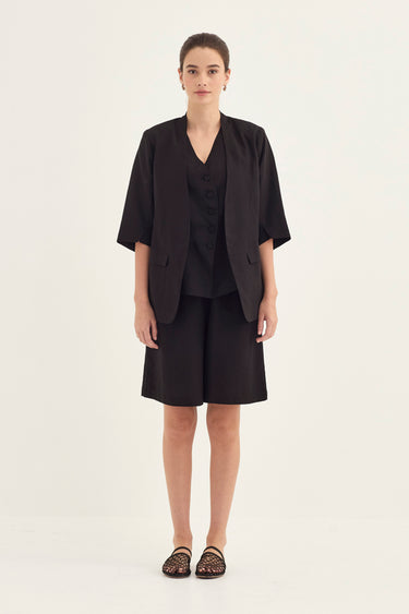 A woman stands against a plain light background wearing a black short-sleeve blazer over a black button-front top and black shorts by Verge, with black strappy flat sandals.
