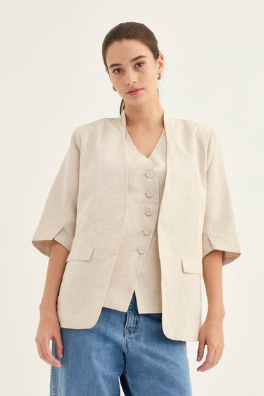 A woman modeling a light beige linen-blend vest with a V-neck and front buttons, styled over a matching beige top and blue jeans by Verge, against a plain off-white studio background.
