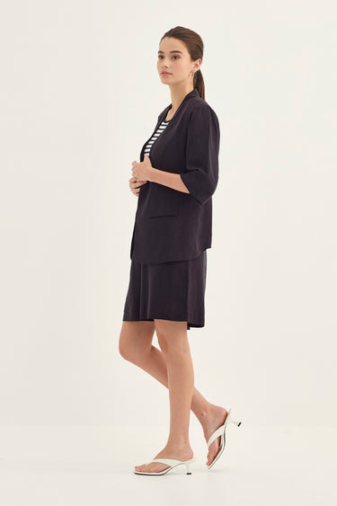 A woman modeling a dark navy blue two-piece set consisting of a short-sleeve blazer and matching skirt, paired with a black and white striped top by Verge and white slide sandals, against a plain off-white studio background.