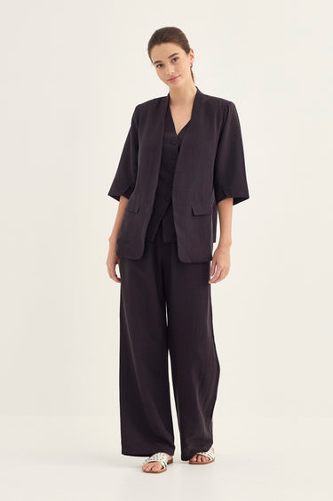 A woman modeling a dark charcoal gray two-piece linen set consisting of a short-sleeve button-front blazer and matching wide-leg trousers by Verge, paired with silver-toned sandals, against a plain off-white studio background.