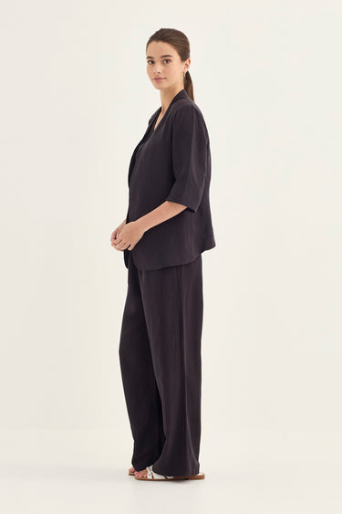 A woman modeling a dark charcoal gray two-piece lounge set consisting of a short-sleeve V-neck top and matching wide-leg pants by Verge, standing against a plain off-white studio background.