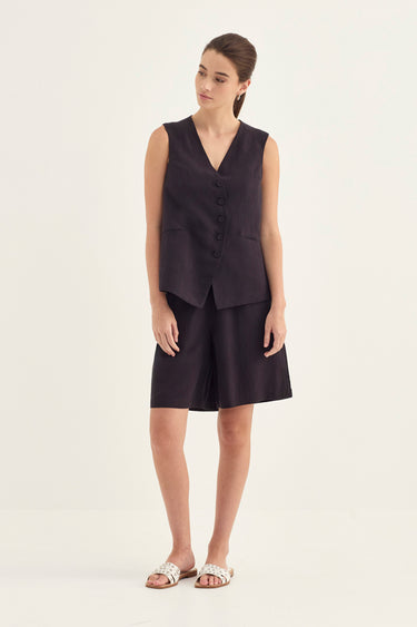 A woman modeling a dark plum sleeveless vest and matching shorts set by Verge, paired with white strappy flat sandals, against a plain off-white studio background.