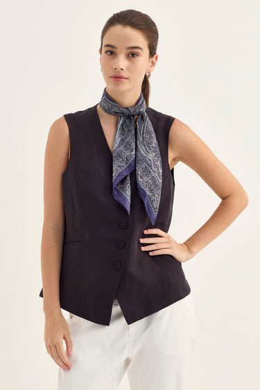 A woman modeling a sleeveless black vest with a patterned blue and purple paisley scarf tied around her neck, paired with white pants by Verge, against a plain off-white background.