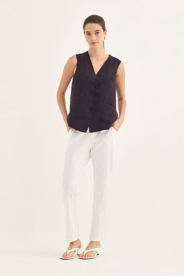 A woman modeling a sleeveless dark plum button-front vest with white trousers by Verge and white sandals, standing against a plain off-white studio background.