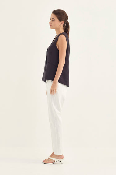 A woman shown in profile wearing a sleeveless dark navy top, white pants, and white sandals by Verge against a plain light background.