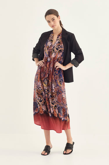 A woman modeling a long-sleeve dress with a complex paisley and floral pattern in red, blue, and gold, styled with a black blazer by Verge and black slide sandals against a plain white studio background.