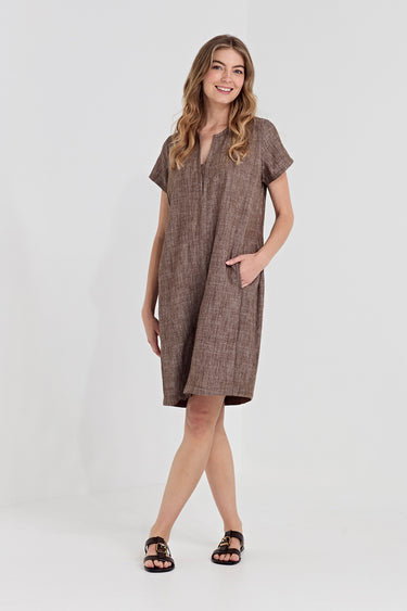 A woman modeling a brown textured linen dress with short sleeves and side pockets by Verge, paired with black sandals, against a plain light gray studio background.