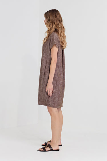 A woman modeling a short-sleeve, knee-length brown textured dress with a subtle plaid pattern by Verge, paired with black slide sandals, shown from a side profile against a light gray studio background.
