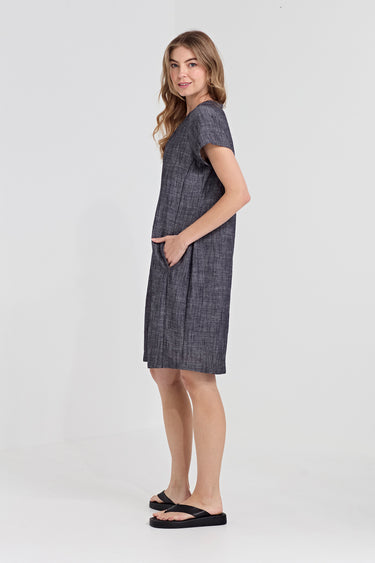 A woman modeling a short-sleeve, knee-length gray textured dress with side pockets by Verge, paired with black flip-flop sandals, against a plain light gray studio background.