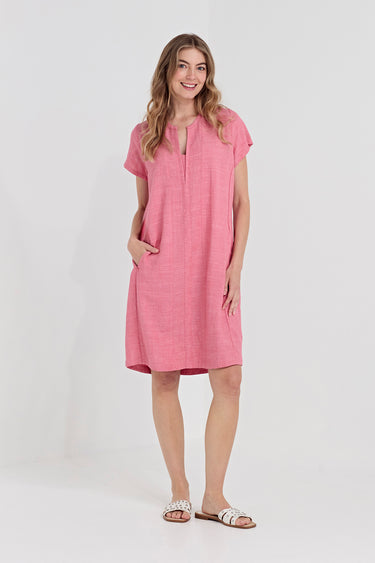 A woman modeling a short-sleeve pink linen dress by Verge with a split neckline and side pockets, standing against a plain white studio background.