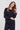 A woman modeling a dark navy blue long-sleeve blouse with button cuffs and matching dark pants by Verge against a light gray studio background.