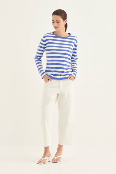 A woman modeling a blue and white horizontally striped long-sleeve knit top with white cropped trousers by Verge and white heeled sandals, standing against a plain off-white studio background.