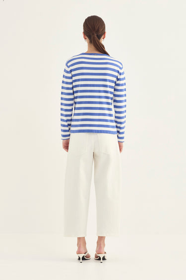 A woman viewed from behind wearing a blue and white striped long-sleeve top, white cropped trousers by Verge, and black-and-white heeled sandals against a plain light background.