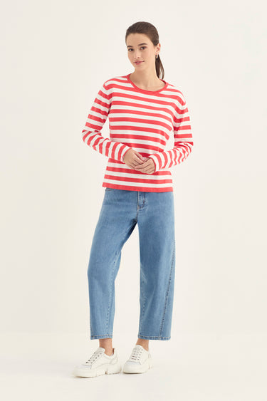 A woman modeling a red and white horizontally striped long-sleeve top, paired with light blue jeans by Verge and white sneakers, against a plain white studio background.