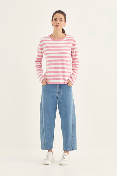 A woman modeling a pink and white horizontally striped long-sleeve knit top with light blue wide-leg jeans by Verge and white sneakers, standing against a plain light gray studio background.