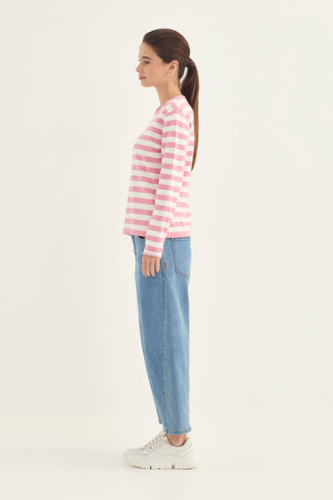 A woman in profile wearing a pink and white striped long-sleeve top, light blue jeans by Verge, and white sneakers, standing against a plain off-white studio background.