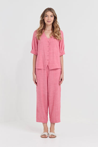 A woman modeling a matching pink two-piece linen set consisting of a short-sleeve button-front top and cropped wide-leg pants by Verge, photographed against a plain light gray studio background.