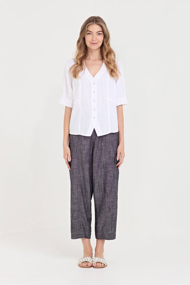 A woman stands against a plain light background wearing a white short-sleeve button-front top and dark gray wide-leg cropped pants by Verge with white embellished sandals.
