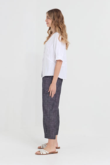 A woman stands in profile wearing a loose white short-sleeve top, dark charcoal plaid cropped pants by Verge, and white slide sandals against a plain light gray studio background.