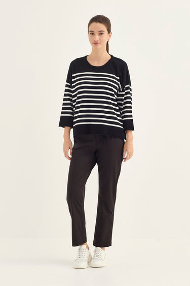 A woman modeling a black and white horizontally striped knit sweater with three-quarter sleeves, paired with black trousers by Verge and white sneakers, against a plain light gray studio background.