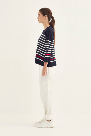 A woman shown in side profile wearing a navy blue knit top with horizontal white and red stripes, white pants by Verge, and white sneakers, photographed against a plain light background.