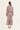 A woman viewed from the back wearing a long-sleeve midi dress with a dusty rose and beige paisley pattern and a matching waist tie by Verge, paired with white heeled sandals against a plain off-white background.