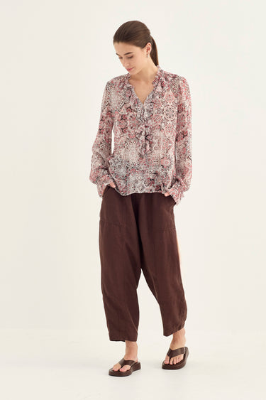 A woman modeling a long-sleeve blouse with a pink, beige, and white paisley pattern, paired with dark brown wide-leg cropped pants and black sandals by Verge, against a plain white studio background.