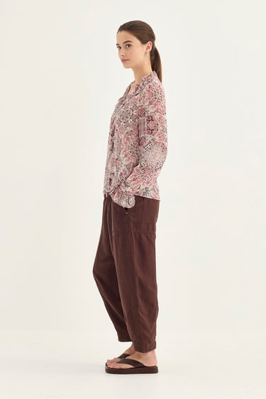 A woman stands in profile wearing a loose, long-sleeve button-up shirt with a muted pink, beige, and white floral pattern, paired with dark brown baggy pants by Verge and black flip-flops, against a plain off-white studio background.