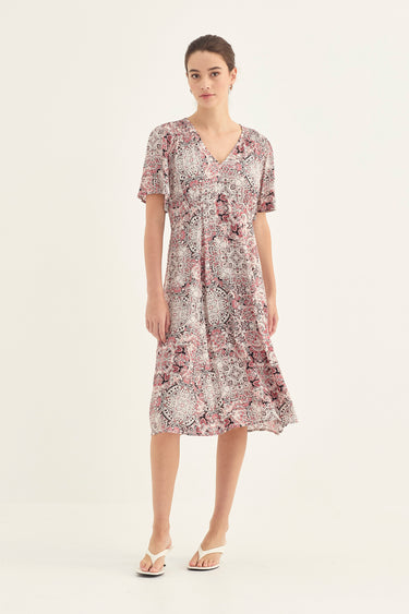 A woman modeling a short-sleeve V-neck midi dress with a dusty pink and white abstract floral print by Verge, paired with white flat sandals, against a plain off-white studio background.