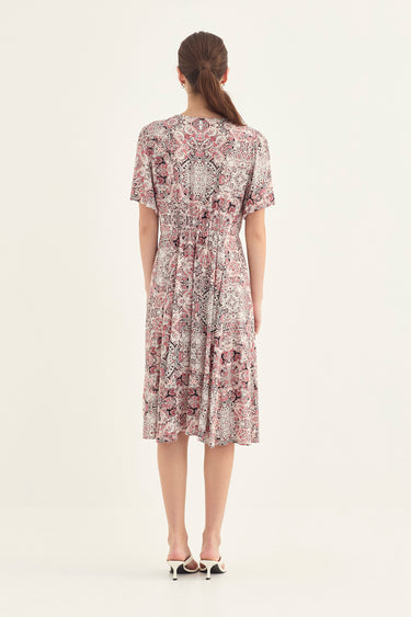 A woman viewed from the back wearing a short-sleeve knee-length dress with an intricate floral and paisley-style print in dusty pink, mauve, and beige tones, paired with white heeled sandals by Verge, against a plain off-white studio background.